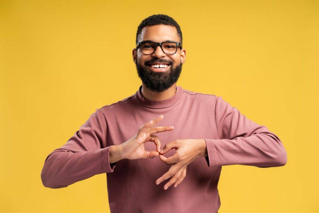 A headshot of a person doing a sign language gesture while standing in front of a yellow background