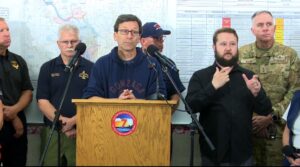 A photo of a white male ASL interpreter with brown hair and a beard interpreting next to Washington State Governor Bob Ferguson at an emergency press conference.